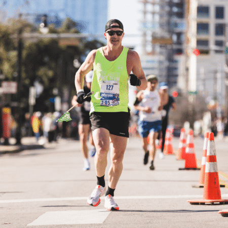 A runner in a neon green tank top, black shorts, and sunglasses competes in the Austin Marathon. He holds a small flag and wears a race bib that reads "Jonathan." Orange cones line the street, with other runners behind him. Austin Marathon Half Marathon & 5K