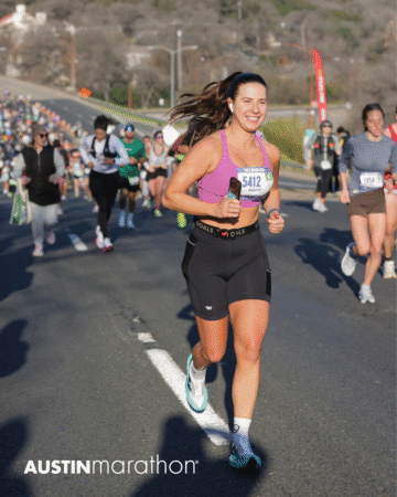 A woman wearing a race bib runs and smiles during the Austin Marathon, with a crowd of other runners behind her on a sunny road. The event logo "AUSTINmarathon" is visible in the bottom left corner, capturing an Official Race Photo. Austin Marathon Half Marathon & 5K