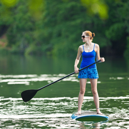 A woman wearing sunglasses, a blue tank top, and blue shorts stands paddleboarding on a calm green lake surrounded by lush trees, enjoying a peaceful day trip in nature. Austin Marathon Half Marathon & 5K