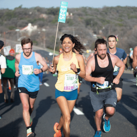 A smiling woman holding a "3:25" race pace flag runs with a group of marathon runners on a sunny Austin Marathon day. Other runners, wearing race bibs, are closely surrounding her on the road. Austin Marathon Half Marathon & 5K