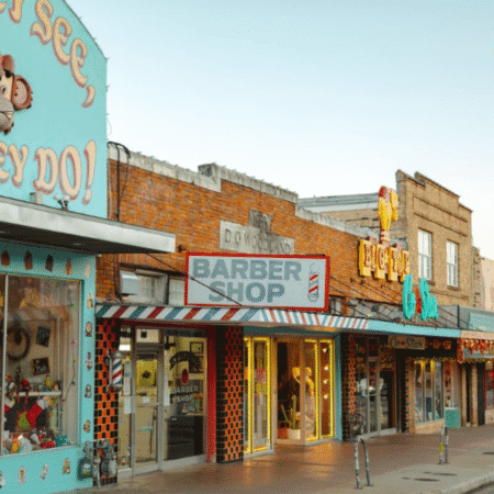 Perfect for a day trip, this colorful street boasts vintage-style shops, including a barber shop with a striped sign and large windows. Unique storefronts feature bright decorations and signage under a clear sky. Austin Marathon Half Marathon & 5K