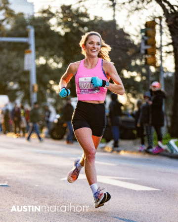 A female runner in a pink top and black shorts smiles while racing in the Austin Marathon, sunlight and spectators behind her. The event logo "AUSTINmarathon" is visible on the street—perfect for your Official Race Photos collection. Austin Marathon Half Marathon & 5K