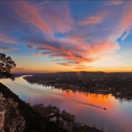 A wide river reflects vibrant pink and orange sunset clouds, perfect for a day trip. A boat creates a wake as it moves down the water, surrounded by trees and houses on the riverbanks. Austin Marathon Half Marathon & 5K