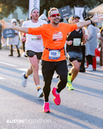 A smiling man in an orange shirt and pink shoes runs with arms outstretched at the Austin Marathon, capturing a personal moment during the race, surrounded by other runners and spectators in the background. Austin Marathon Half Marathon & 5K