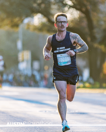 A male runner wearing a black tank top and glasses races in the Austin Marathon, race bib number 10006 visible. Sunlight and trees provide a scenic backdrop for this personal moment captured in official race photos. Austin Marathon Half Marathon & 5K
