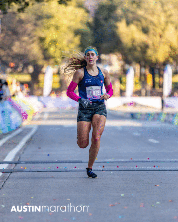 A female runner with a race bib and pink arm sleeves races down a city street during the Austin Marathon, with blurred trees and spectators in the background. The image captures an exciting personal moment from the Official Race Photos collection. Austin Marathon Half Marathon & 5K
