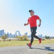 A man in a red shirt, black leggings, and a cap jogs on a paved path in a park on a sunny day, following his beginner runner checklist, with a city skyline and green grass in the background. Austin Marathon Half Marathon & 5K