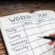 An open notebook with a grid pattern displays a handwritten, two-week workout plan, perfect for a beginner runner. Days of the week are listed, and a hand holding a pen is poised over the page, ready to fill in this runner checklist. Austin Marathon Half Marathon & 5K