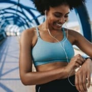 Woman in athletic wear smiles and checks her smartwatch on a blue pedestrian bridge. Earphones in, she enjoys music during her workout—perfect for any beginner runner following their runner checklist. Austin Marathon Half Marathon & 5K