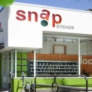 A modern storefront with large windows and the sign "snap KITCHEN" in red and green letters above the entrance, this spot stands out among healthy Austin restaurants. The building exterior is white with green accents. Austin Marathon Half Marathon & 5K