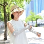 A woman wearing a wide-brimmed hat and sleeveless white top smiles while putting on long, light gray arm sleeves outdoors—a picture-perfect moment for any beginner runner enjoying a sunny day, with green trees and tall buildings in the background. Austin Marathon Half Marathon & 5K