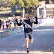 A male runner in athletic gear crosses a finish line with arms raised, smiling, as colorful confetti falls around him—an inspiring moment for any beginner runner at a festive race event filled with cheering spectators. Austin Marathon Half Marathon & 5K