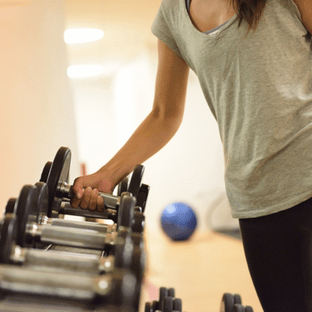 A person wearing a gray t-shirt and black pants picks up a dumbbell from a rack in a gym, focusing on strength training for runners. Exercise equipment and a blurred blue yoga ball are visible in the background. Austin Marathon Half Marathon & 5K
