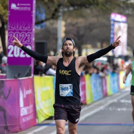 A male runner in a black tank top and headband crosses the marathon finish line with arms raised in victory, fueled by marathon motivation. Spectators cheer along colorful barricades, while event banners mark the finish area. Austin Marathon Half Marathon & 5K