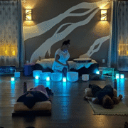 A woman plays crystal singing bowls in a dimly lit Austin room with glowing blue lights. Two people enjoy post-run recovery on mats and cushions, as candles and soft wall art create a calming atmosphere—a hidden gem for relaxation. Austin Marathon Half Marathon & 5K
