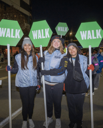 Three women stand side by side at night, each holding a green octagonal "WALK" sign. Dressed warmly and smiling, they embrace volunteer opportunities, with other people and city lights glowing in the background. Austin Marathon Half Marathon & 5K