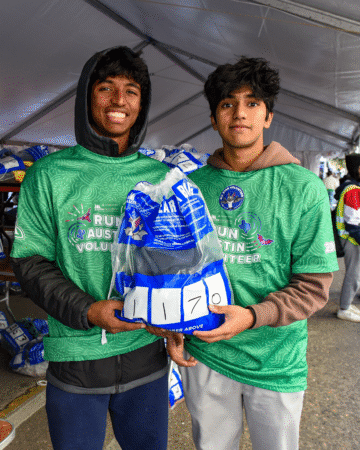 Two young men wearing green volunteer shirts hold a clear bag labeled "1170" under a tent at an outdoor event, smiling at the camera. The scene highlights volunteer opportunities, with others and bags visible in the background. Austin Marathon Half Marathon & 5K