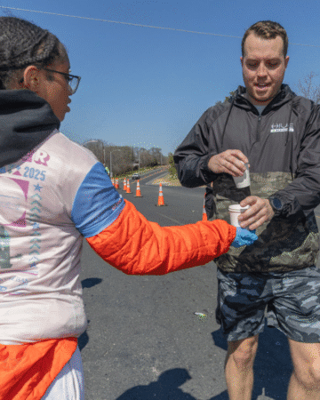 A volunteer explores new volunteer opportunities by handing a cup of water to a runner during a race on a sunny day, with orange cones and a clear blue sky in the background. Austin Marathon Half Marathon & 5K