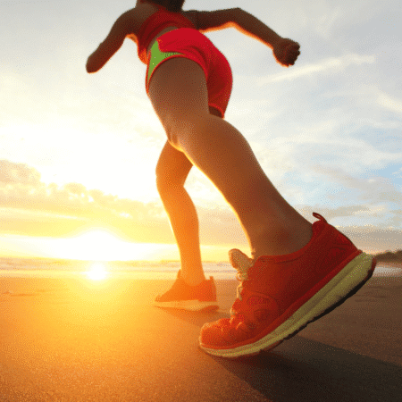 A person in red athletic shoes and shorts jogs along a beach at sunrise, the sun casting a warm glow on the sand—a perfect scene for running training or tempo runs by the shore. Austin Marathon Half Marathon & 5K