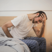A man in a white t-shirt and plaid pajama pants sits on a bed, leaning forward with one hand on his forehead and a pained expression, capturing the struggle to overcome a training slump. Austin Marathon Half Marathon & 5K