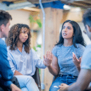 Four people sit in a circle having a serious discussion. One woman gestures expressively about overcoming a training slump, while another listens attentively with a supportive hand on her arm. The setting appears informal and indoors. Austin Marathon Half Marathon & 5K