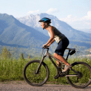 A woman wearing a helmet rides a mountain bike on a gravel path, surrounded by green grass and wildflowers, with tall mountains and a blue sky in the background—embracing injury prevention strategies for the long haul. Austin Marathon Half Marathon & 5K
