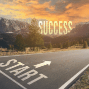 A paved road with "START" and an arrow painted on it leads toward mountains at sunset. "SUCCESS" appears in large letters in the sky ahead, inspiring the runner’s mental reset and symbolizing a journey fueled by setting mini goals. Austin Marathon Half Marathon & 5K