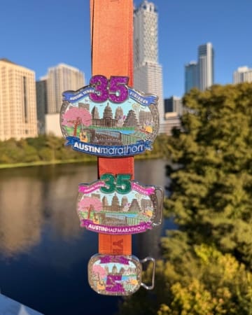 Three colorful marathon medals hang on an orange ribbon for the 35th Austin Marathon, Half Marathon, and 5K—perfect race bling—with a city skyline and river in the background on a sunny day. Austin Marathon Half Marathon & 5K Three colorful marathon medals hang on an orange ribbon for the 35th Austin Marathon, Half Marathon, and 5K—perfect race bling—with a city skyline and river in the background on a sunny day. Austin Marathon Half Marathon & 5K