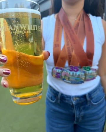 A person wearing a white tank top, blue jeans, and colorful marathon medals holds a pint of beer toward the camera. The outdoor scene suggests a sunny celebration filled with race bling and accomplishment. Austin Marathon Half Marathon & 5K A person wearing a white tank top, blue jeans, and colorful marathon medals holds a pint of beer toward the camera. The outdoor scene suggests a sunny celebration filled with race bling and accomplishment. Austin Marathon Half Marathon & 5K