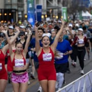 Three smiling women in red athletic outfits with race bibs cheer and raise their arms while running together as a team in a large outdoor charity race; many other runners are behind them on the city street. Austin Marathon Half Marathon & 5K