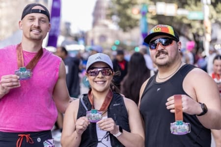 Three runners smile and hold up medals after a race, representing their community teams. They stand outdoors with a crowd and a domed building in the blurred background, wearing athletic gear, sunglasses, and hats. Austin Marathon Half Marathon & 5K Three runners smile and hold up medals after a race, representing their community teams. They stand outdoors with a crowd and a domed building in the blurred background, wearing athletic gear, sunglasses, and hats. Austin Marathon Half Marathon & 5K