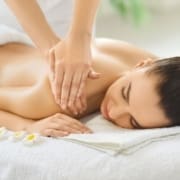 A woman lies face down on a white towel, smiling with her eyes closed as she receives a relaxing shoulder massage—perfect for physical and mental recovery. Frangipani flowers are placed beside her on the towel. Austin Marathon Half Marathon & 5K