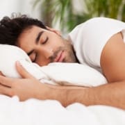 A man with dark hair is sleeping peacefully on a white bed, hugging a pillow. He is wearing a white shirt, and there are green plants blurred in the background, highlighting the importance of physical recovery after intense activity. Austin Marathon Half Marathon & 5K