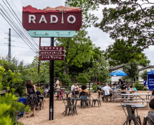 Outdoor patio spot with people sitting at tables under trees. A large vintage "RADIO" sign and smaller signs advertise coffee, cocktails, and live music. It’s a casual, relaxed hangout—perfect for runners enjoying Spring in Austin. Austin Marathon Half Marathon & 5K