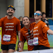 Three smiling runners in matching orange shirts reading "I THOUGHT THIS WAS A BAR CRAWL?" pose happily at a race event, looking like school teams or collegiate teams with their arms around each other and race bibs proudly displayed. Austin Marathon Half Marathon & 5K