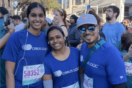 Three people wearing matching blue "Cirrus Logic" shirts smile at the camera during a race event, representing Corporate Teams. Two have race bibs pinned to their shirts, surrounded by other participants in an outdoor city setting. Austin Marathon Half Marathon & 5K Three people wearing matching blue "Cirrus Logic" shirts smile at the camera during a race event, representing Corporate Teams. Two have race bibs pinned to their shirts, surrounded by other participants in an outdoor city setting. Austin Marathon Half Marathon & 5K