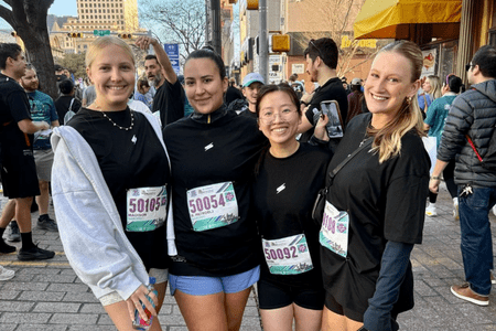 Four women wearing black shirts and race bibs smile and pose together at an outdoor race event—perfect for team building. Other participants and spectators are visible in the background on a bustling city street. Austin Marathon Half Marathon & 5K Four women wearing black shirts and race bibs smile and pose together at an outdoor race event—perfect for team building. Other participants and spectators are visible in the background on a bustling city street. Austin Marathon Half Marathon & 5K