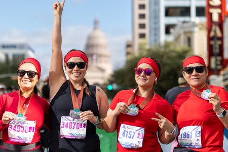 Four women wearing red shirts and headbands proudly display medals after a charity race, smiling for the camera. One woman raises her arm in celebration. City buildings and the Texas State Capitol are visible in the background. Austin Marathon Half Marathon & 5K Four women wearing red shirts and headbands proudly display medals after a charity race, smiling for the camera. One woman raises her arm in celebration. City buildings and the Texas State Capitol are visible in the background. Austin Marathon Half Marathon & 5K