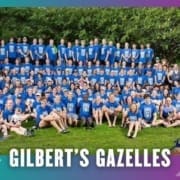 A large group of people wearing matching blue shirts pose for a group photo on grass with trees in the background, representing their running club. Text at the bottom reads "GILBERT'S GAZELLES" with a small gazelle logo on the right. Austin Marathon Half Marathon & 5K