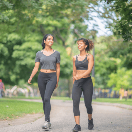 Two women in athletic wear walk and smile together on a tree-lined park path, sharing running motivation and celebrating their next big goal on a sunny, greenery-filled day. Austin Marathon Half Marathon & 5K