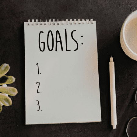 A spiral notebook with "GOALS:" at the top and numbers 1, 2, and 3 on a blank page—perfect for jotting down your next big goal—sits beside a white pen, plant, and cup on a dark surface. Austin Marathon Half Marathon & 5K
