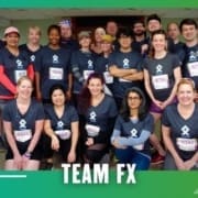 A group of people wearing matching "TEAM FX" shirts and race bibs pose together indoors, smiling, in front of a green and blue graphic border with "TEAM FX" text at the bottom, celebrating their Group Run. Austin Marathon Half Marathon & 5K