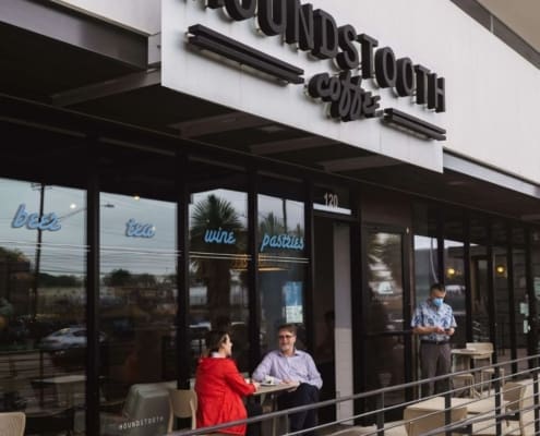 Two people sit at an outdoor table in front of Houndstooth Coffee, one of the best coffee shops Austin offers. A person in a mask stands nearby while the window advertises "best tea wine pastries." Ideal spot for post-run meetups. Austin Marathon Half Marathon & 5K