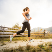 A woman in athletic clothing runs outdoors on a dirt path beside a wooden fence, mountains rising in the background—a reminder to keep pushing forward, even when motivation fades beneath a cloudy sky. Austin Marathon Half Marathon & 5K