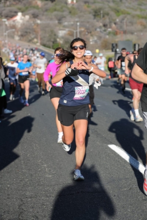 A woman running in a road race smiles and forms a heart with her hands, surrounded by other runners. Sporting a navy shirt and bib, she enjoys the sun—perhaps fueled by Austin Marathon hill training or practicing how to run hills efficiently. Austin Marathon Half Marathon & 5K