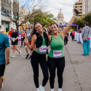Two women in athletic wear smile and pose with peace signs after finishing a race, proving that even when motivation fades, determination carries them forward. Other runners surround them on a city street with the Texas State Capitol in the background. Austin Marathon Half Marathon & 5K