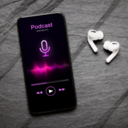 A smartphone displaying a podcast episode screen with a microphone icon and sound waves, next to white wireless earbuds on a dark stone surface—perfect for listening when motivation fades. Austin Marathon Half Marathon & 5K