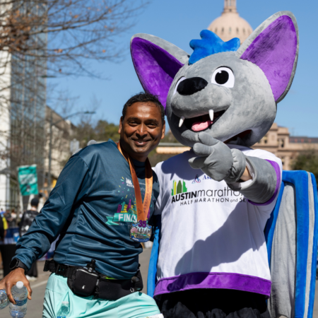 A smiling marathon finisher poses with a large bat mascot in an "Austin Marathon" shirt, with the Texas State Capitol building in the background—a reminder to celebrate achievements even when motivation fades. The runner proudly wears a medal and race gear. Austin Marathon Half Marathon & 5K
