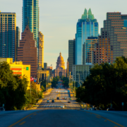 A view of downtown Austin, Texas, with the Texas State Capitol framed by skyscrapers—an iconic scene along an Austin bucket-list marathon route, seen from a wide tree-lined street in the glow of early morning sunlight. Austin Marathon Half Marathon & 5K