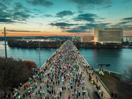 A large crowd of runners crosses a bridge over a river at sunrise or sunset, with tall buildings on either side and a colorful sky in the background—perfect scenery for Austin Marathon hill training or running uphill training workouts. Austin Marathon Half Marathon & 5K